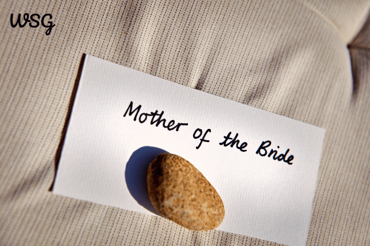 Close-up of a 'Mother of the Bride' place card held down by a stone, placed on a textured beige fabric, used for wedding seating arrangements