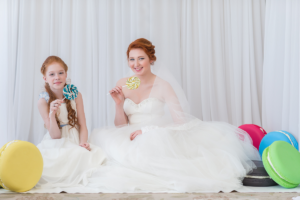 two sisters wearing cute wedding dresses sitting beside each other holding lollipops.