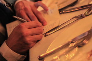 Person writing a short wedding speech at a dinner table setting.